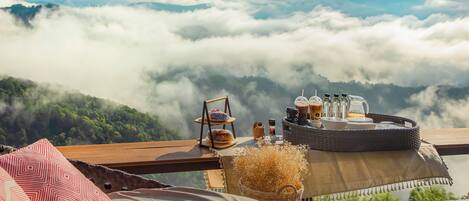 A cozy outdoor seating area with a table set for breakfast, overlooking a mountainous landscape with clouds below.