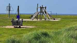 A playground with a slide and climbing structures, set against a grassy field with a clear sky.