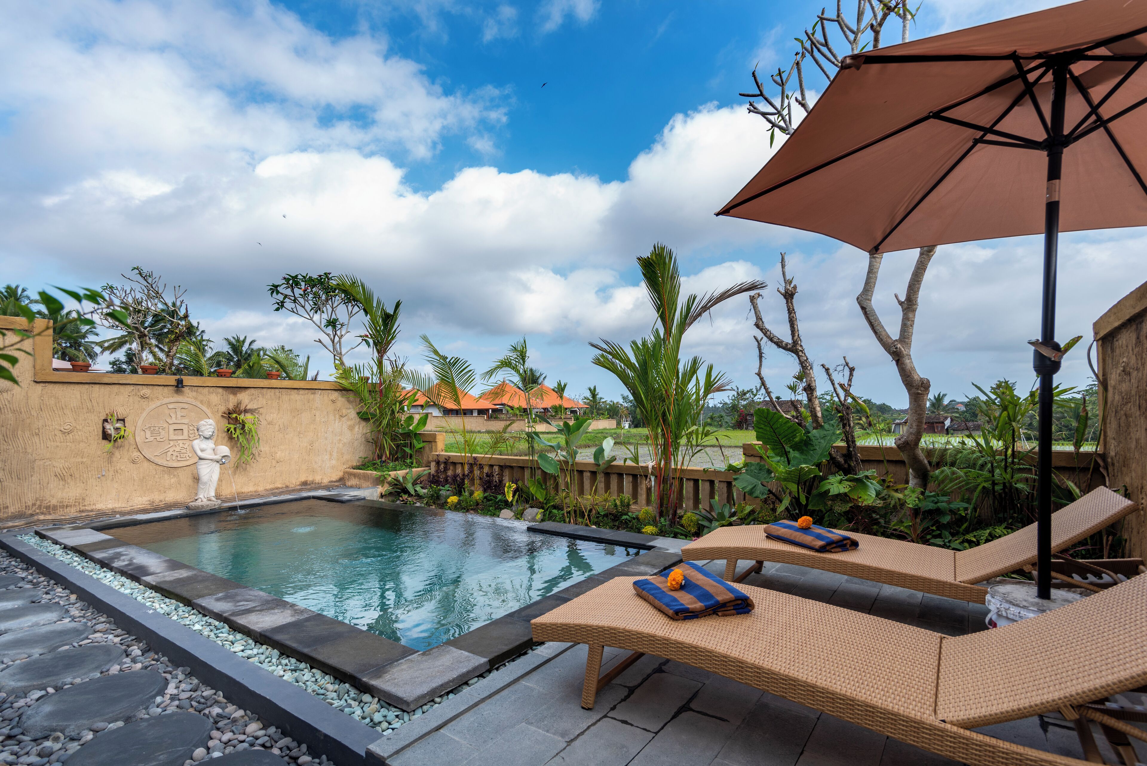 A pool area with lounge chairs and an umbrella, surrounded by tropical plants and a stone pathway.