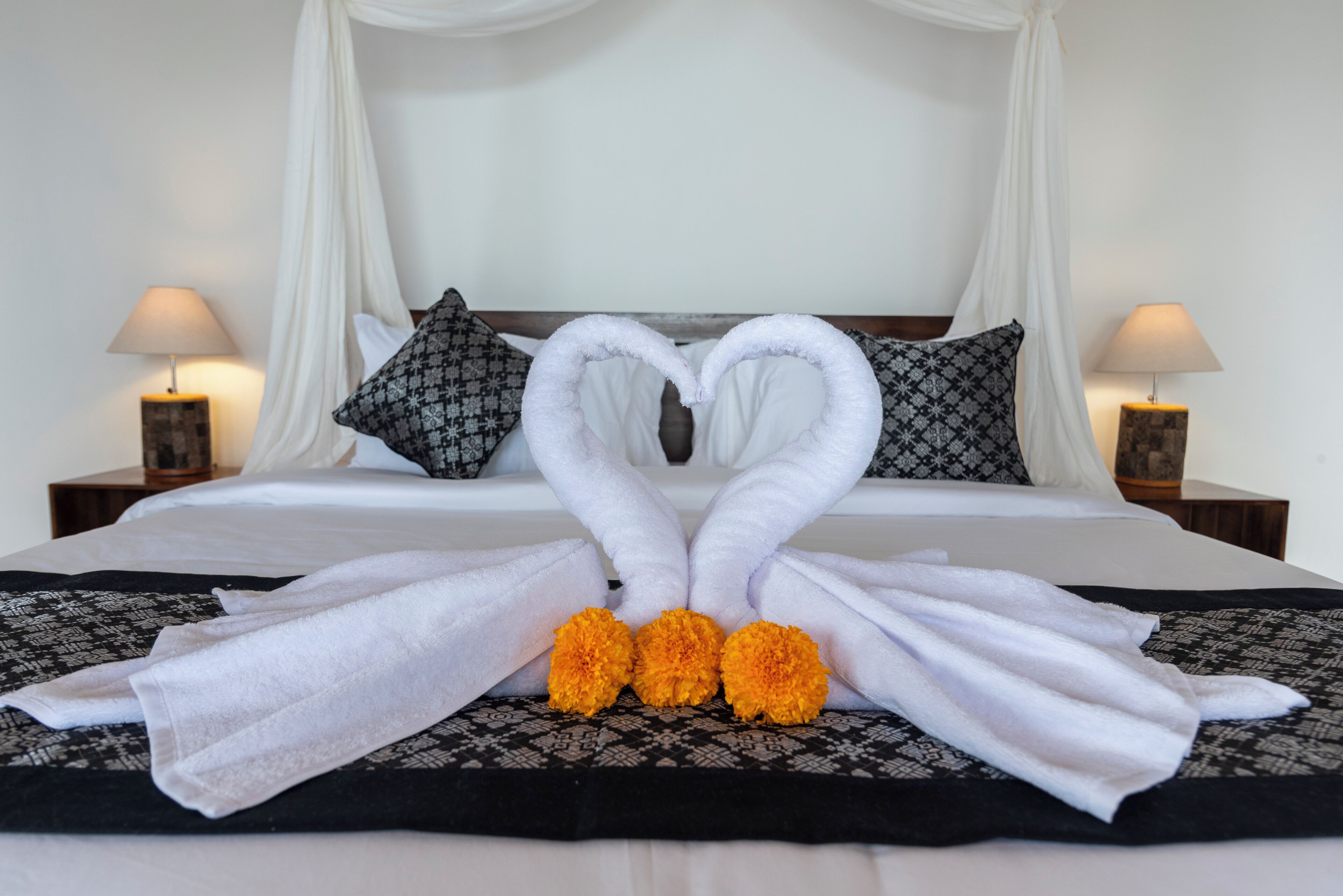 A neatly made bed with white and black patterned bedding, a heart-shaped towel arrangement, and two nightstands with lamps.