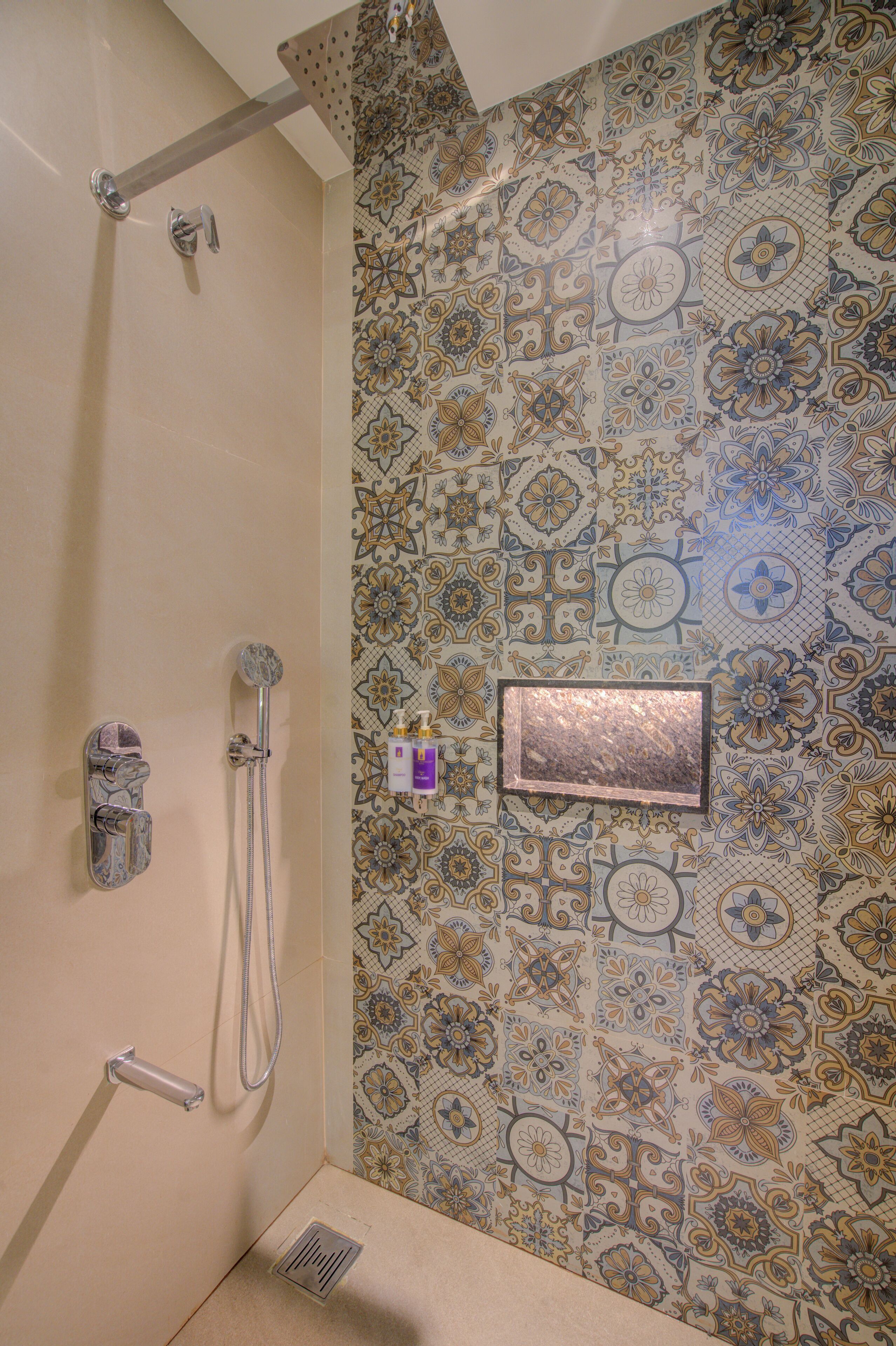 A shower with a handheld showerhead and a wall-mounted faucet. The shower area has a tiled wall with a floral pattern.