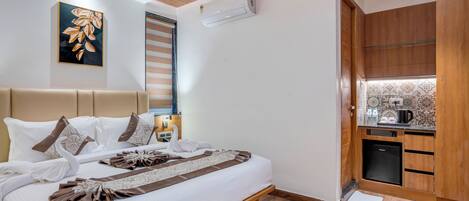 A hotel room with a bed, a ceiling fan, an air conditioner, a small kitchen area, and a decorative wall piece.