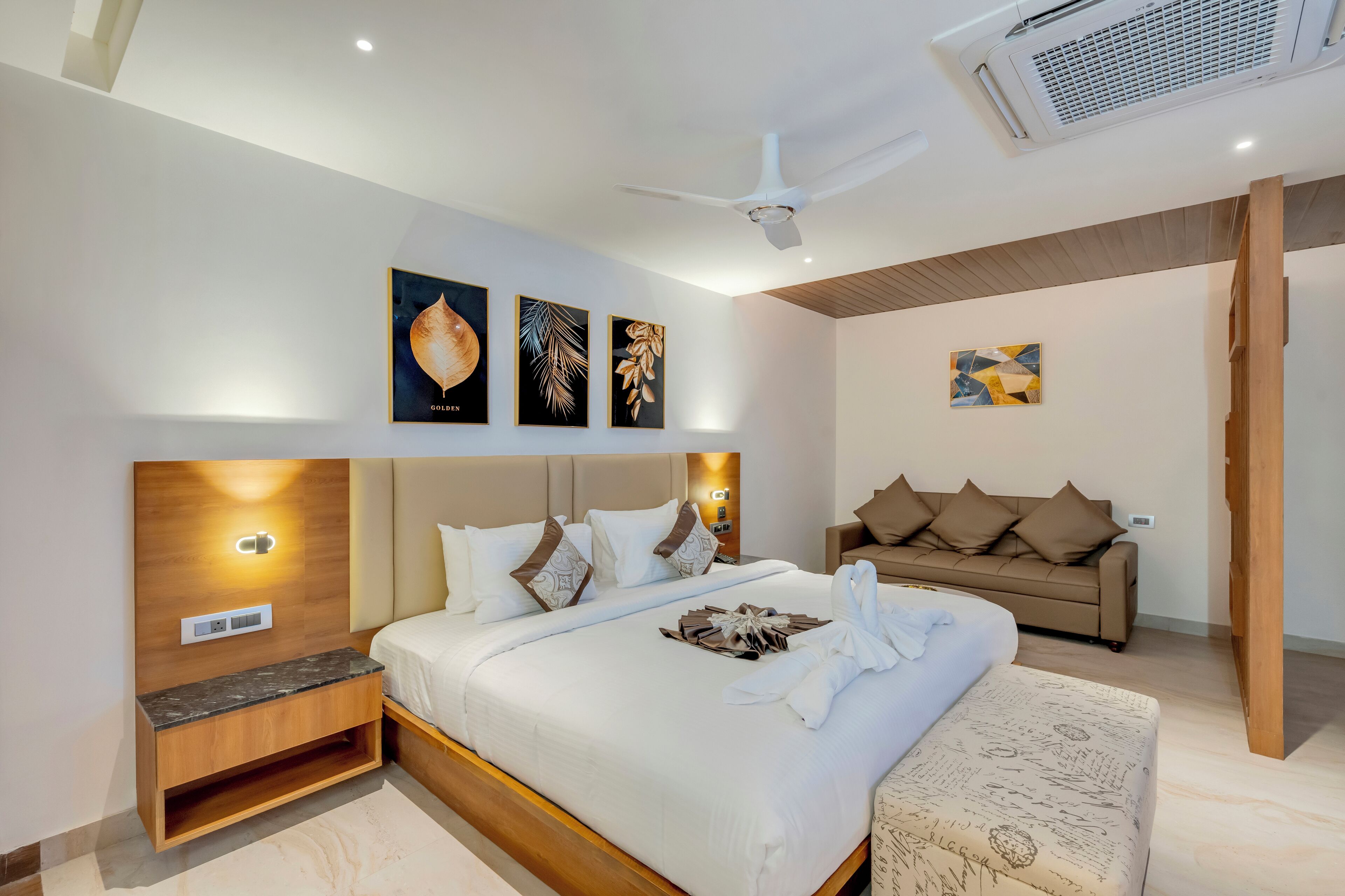 A modern hotel room with a large bed, a wooden headboard, a sofa, and a ceiling fan.