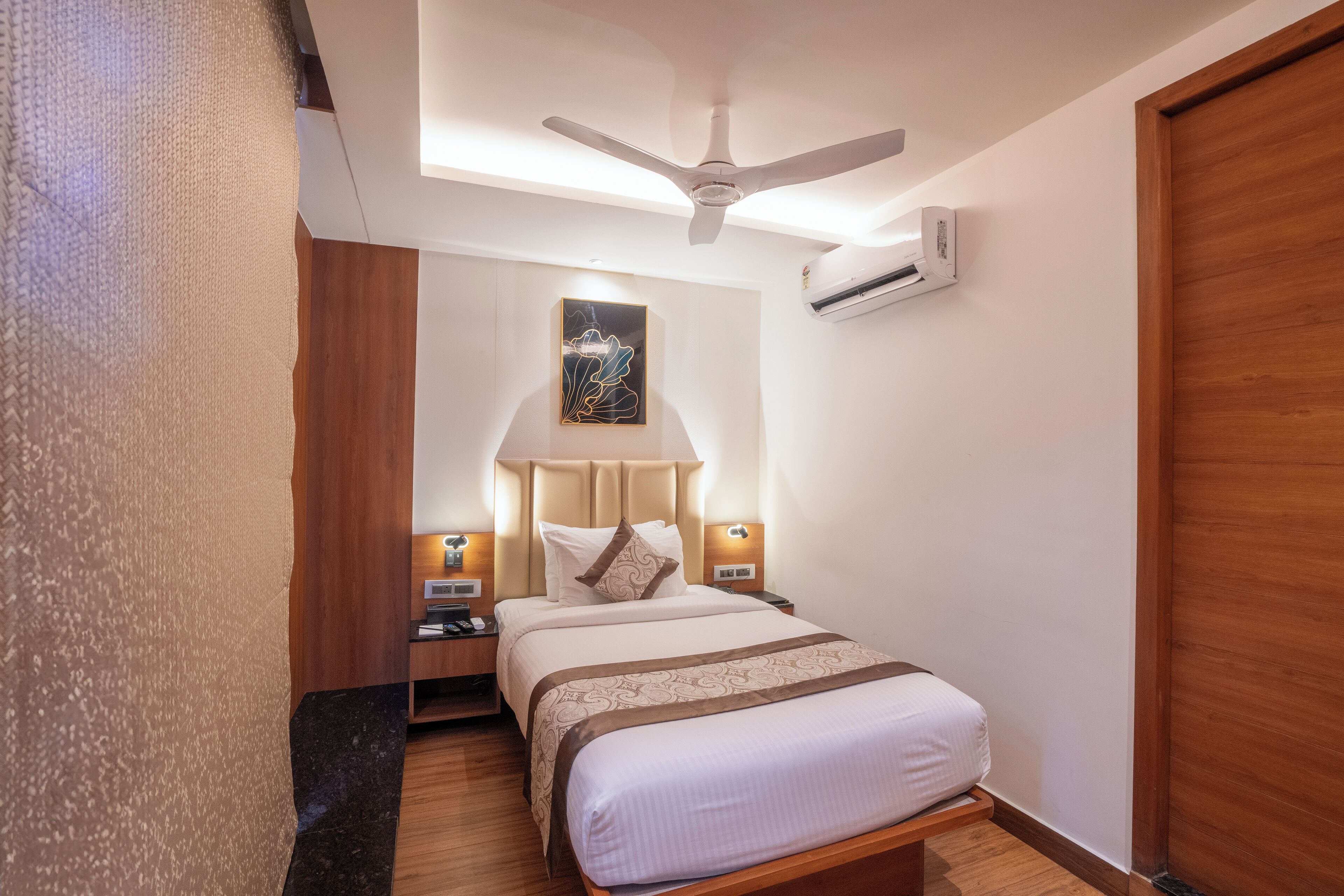 Una habitación de hotel con cama, ventilador de techo, unidad de aire acondicionado, mesita de noche, lámpara y una obra de arte colgada en la pared.