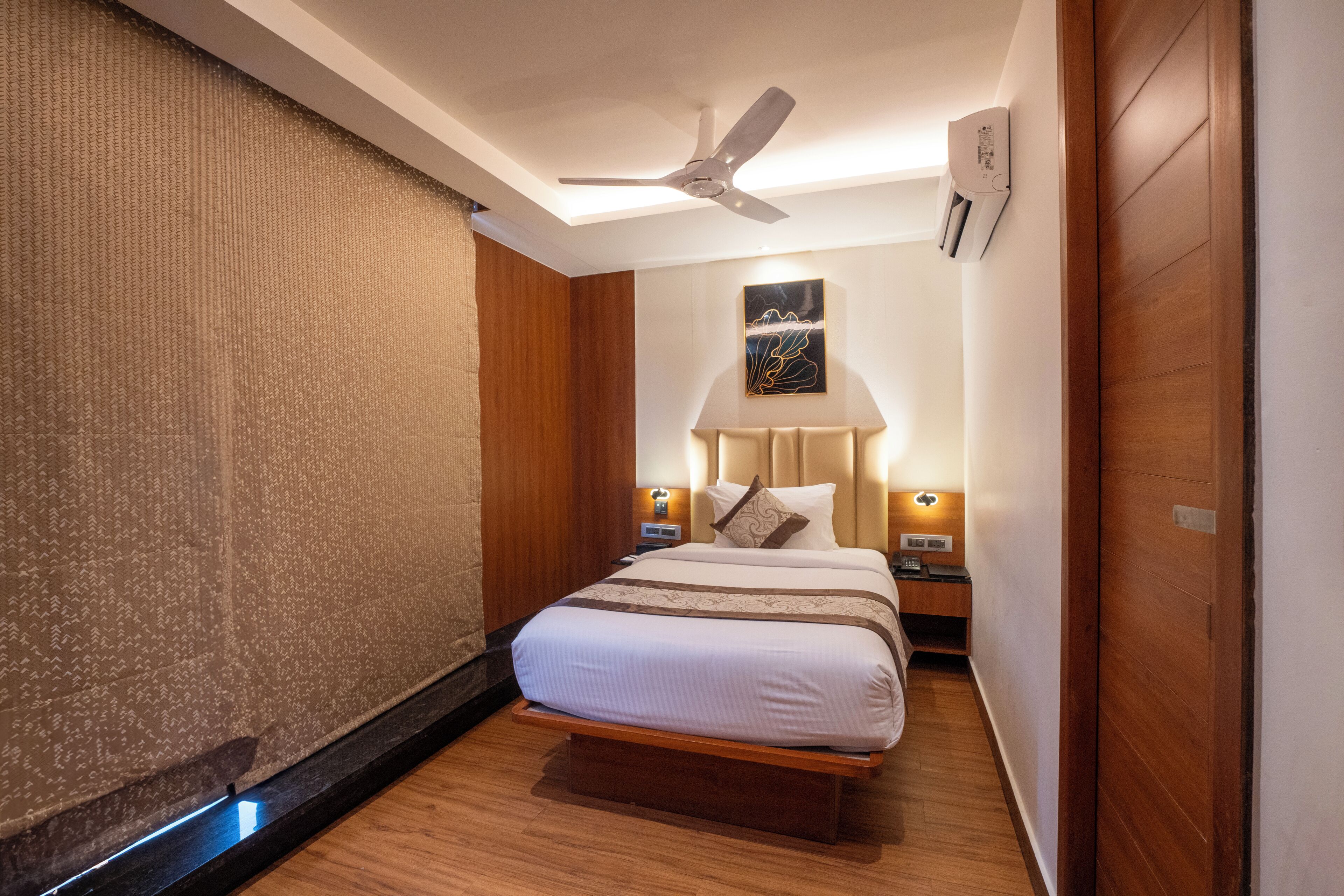 A hotel room with a bed, a ceiling fan, a wall-mounted air conditioner, and a nightstand with a lamp.