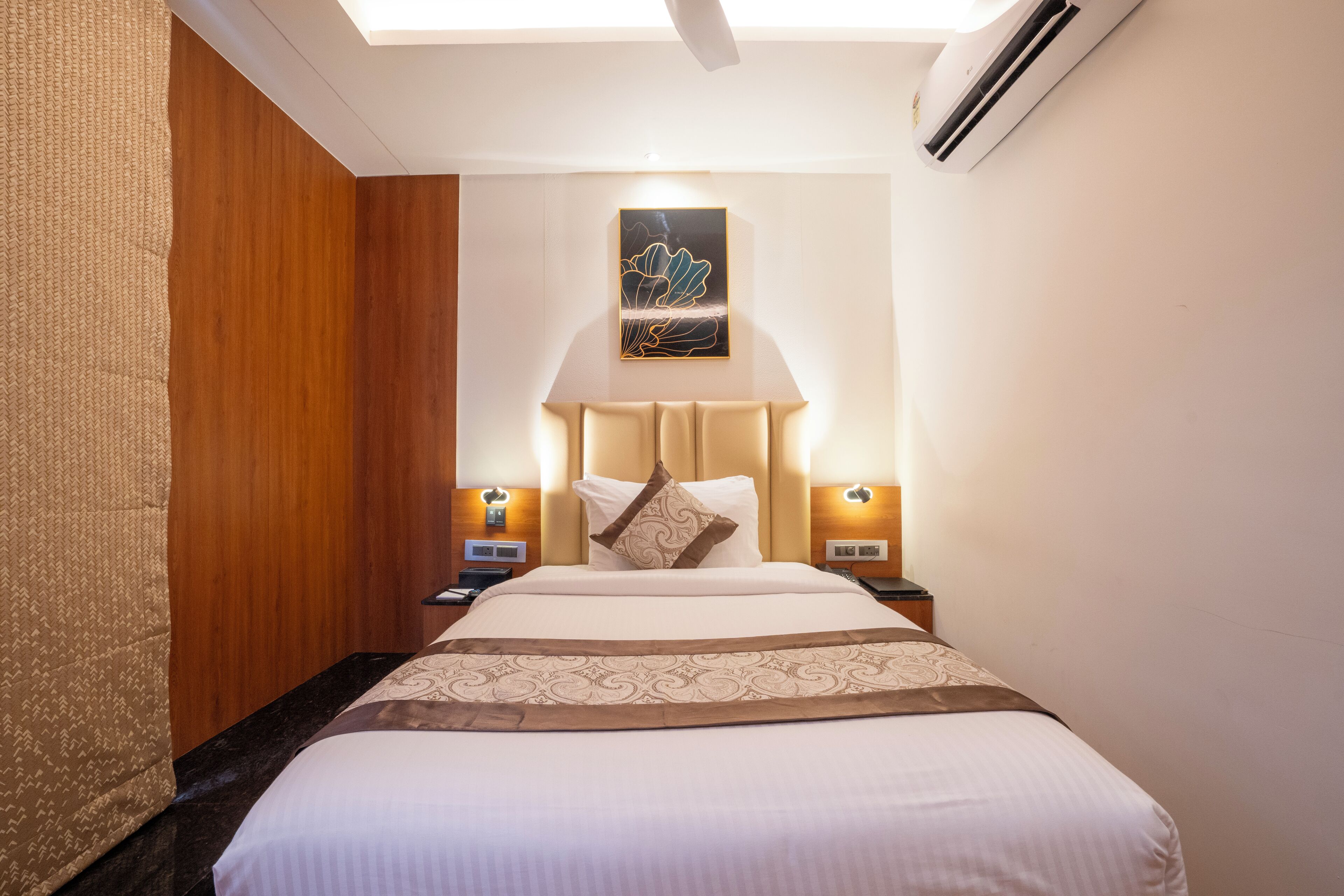 A hotel room with a large bed, two bedside tables, a wall-mounted air conditioner, and a ceiling light.