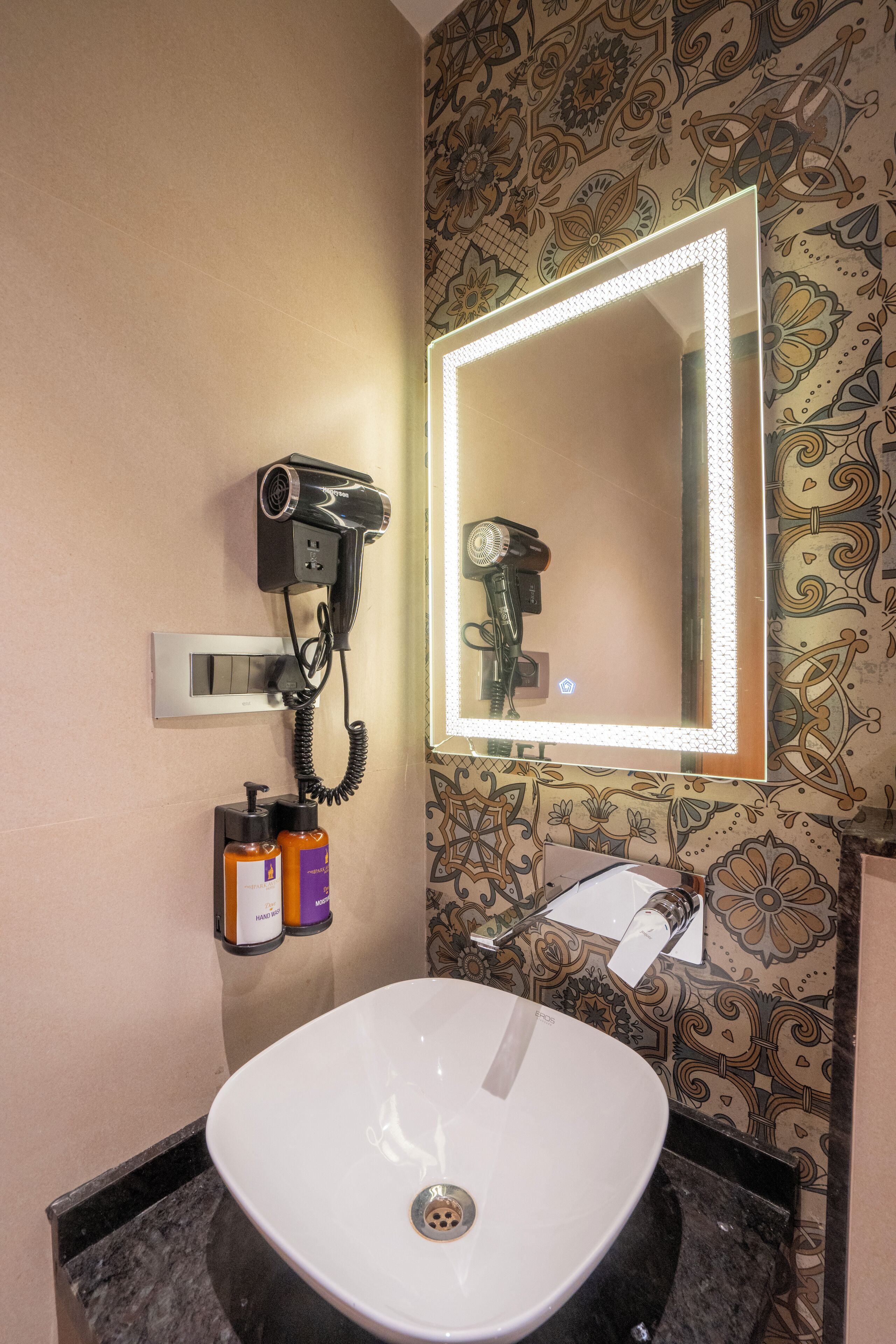 Ein Badezimmer mit weißem Waschbecken, einem an der Wand montierten Föhn und einem Spiegel mit LED-Beleuchtung.