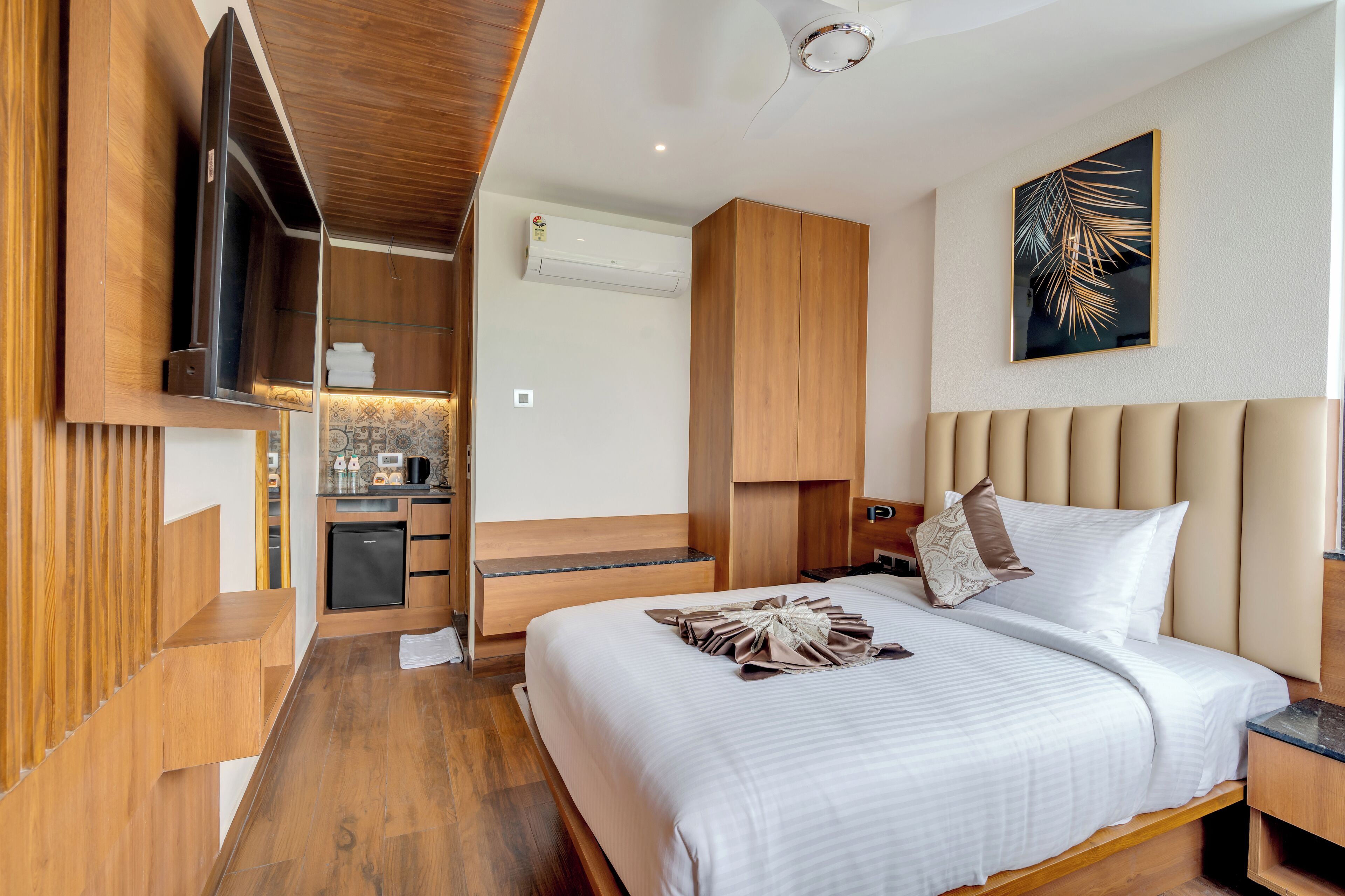 A modern hotel room with a large bed, wooden furniture, a flat-screen TV, and a small kitchenette.
