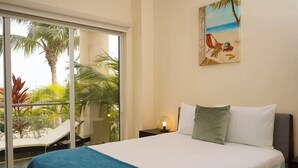 Hotelkamer met een bed, nachtkastje met een lamp, schilderij van een strandtafereel, en een schuifdeur die leidt naar een balkon met een stoel en palmen.