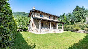 A stone house with a covered porch, surrounded by a well-maintained lawn and trees. - Villa Vittoriano - Surrounded By Nature (Camaiore)