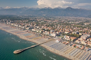 An aerial view of a coastal city with a long pier extending into the sea, a densely packed urban area, and mountains in the background.
