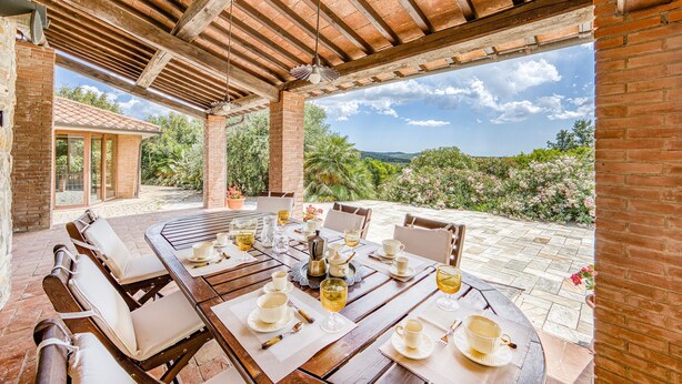 Un patio al aire libre con una mesa de madera dispuesta para comer, rodeado de muros de ladrillo y vistas a zonas verdes y colinas.