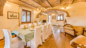 Un comedor con una mesa larga dispuesta para una comida, rodeada de sillas blancas, un sofá y un piso de madera.