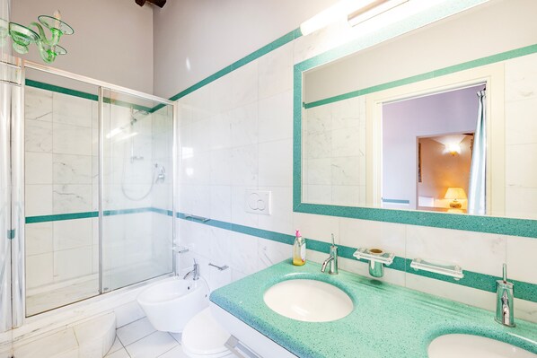 Un baño con ducha envuelta en cristal, dos lavamanos y un espejo grande con detalles color turquesa.