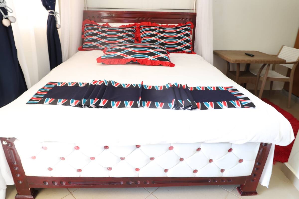 A neatly made bed with a patterned bedspread and pillows, a wooden headboard, a small table, and a chair in a hotel room.