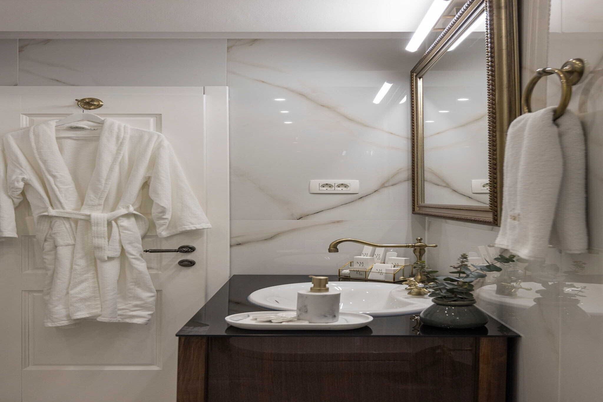 Un baño con pared de mármol, un espejo grande, un lavamanos con grifo dorado y una bata colgada en la puerta.