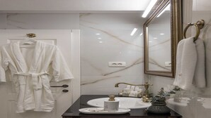 Un baño con pared de mármol, un espejo grande, un lavamanos con grifo dorado y una bata colgada en la puerta.