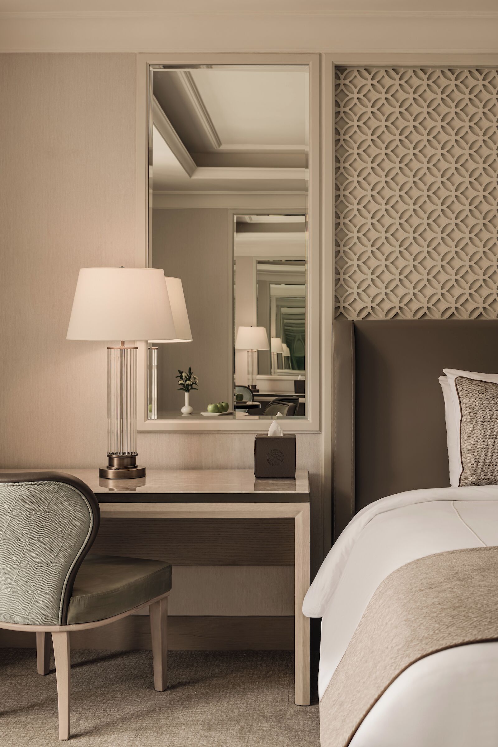 A hotel room with a bed, a desk, a chair, a lamp, a mirror, and a decorative wall panel.