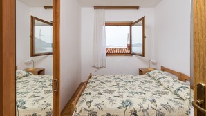 A bedroom with two beds, a window with a view of a red-roofed building, and a mirror reflecting the room.