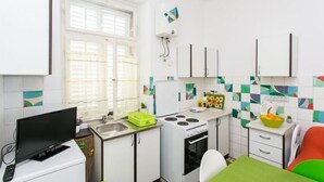 Een kleine keuken met witte kastjes, een fornuis, een spoelbak en een raam met jaloezieën.