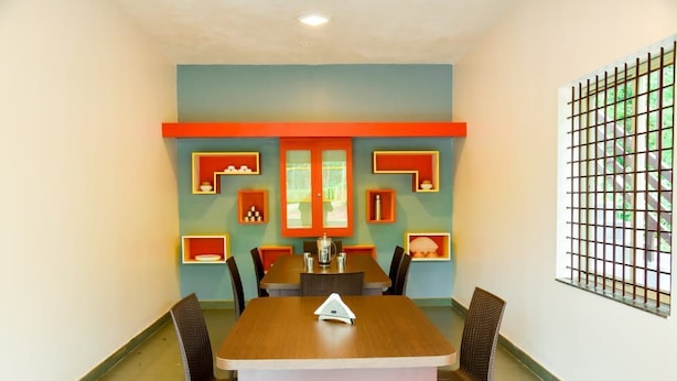 A dining area with a wooden table and wicker chairs, a red accent wall, and a window with a metal grid. - Nature Kingdom (Chikkamagaluru)
