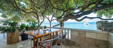 An outdoor dining area with a table and chairs, surrounded by potted plants and overlooking a body of water with a boat in the distance.