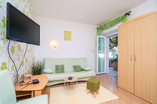 Una sala de estar con televisor de pantalla plana, un sofá, una mesita redonda pequeña, un mueble y una cortina verde.