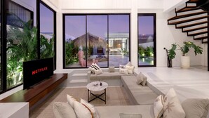 A modern living room with a large TV, comfortable seating, and large windows offering a view of a pool and outdoor area. - Aquamarine Villas III (Munggu)