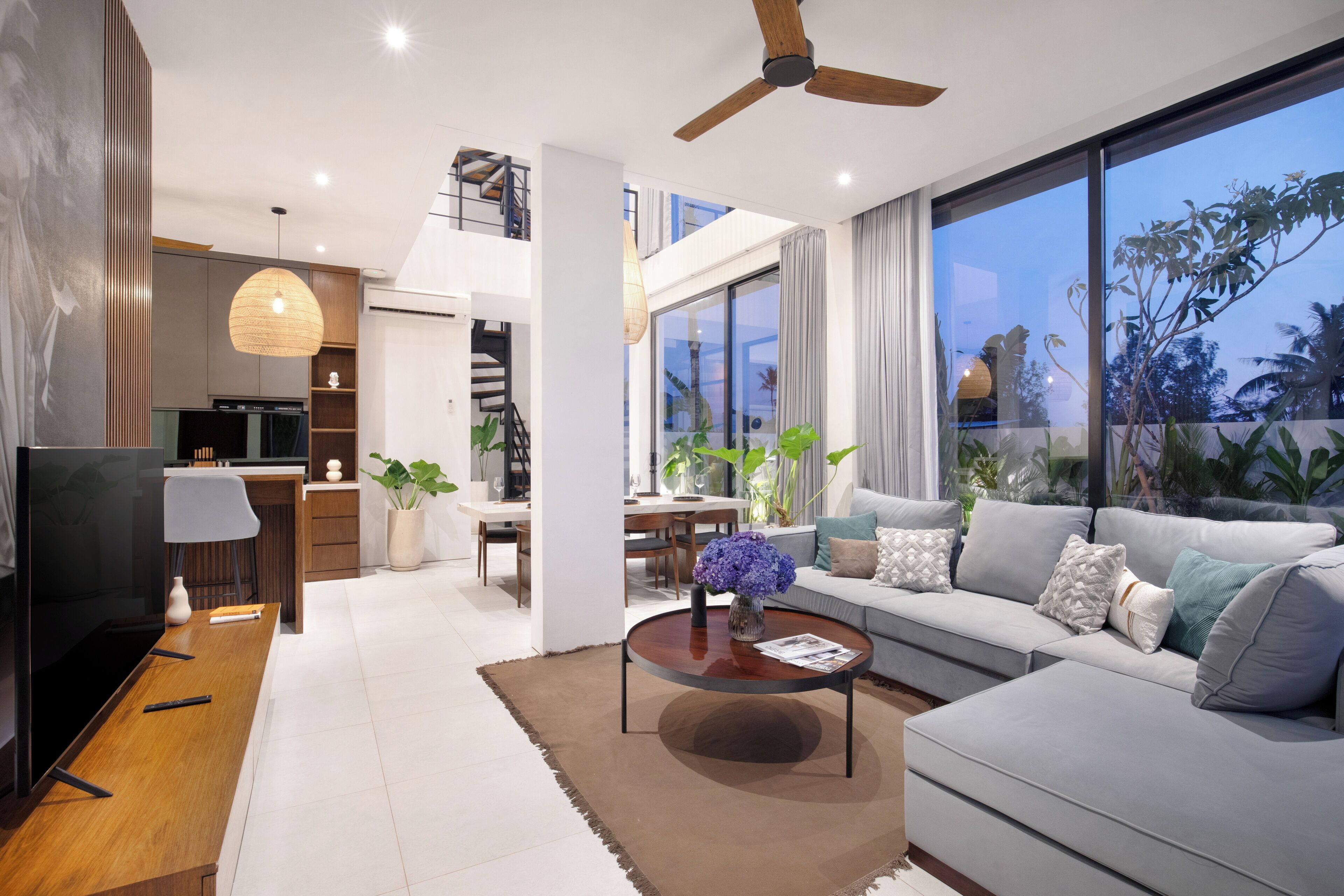 A modern living room with a sofa, coffee table, and TV. There is a dining area with a table and chairs, and a kitchen in the background.