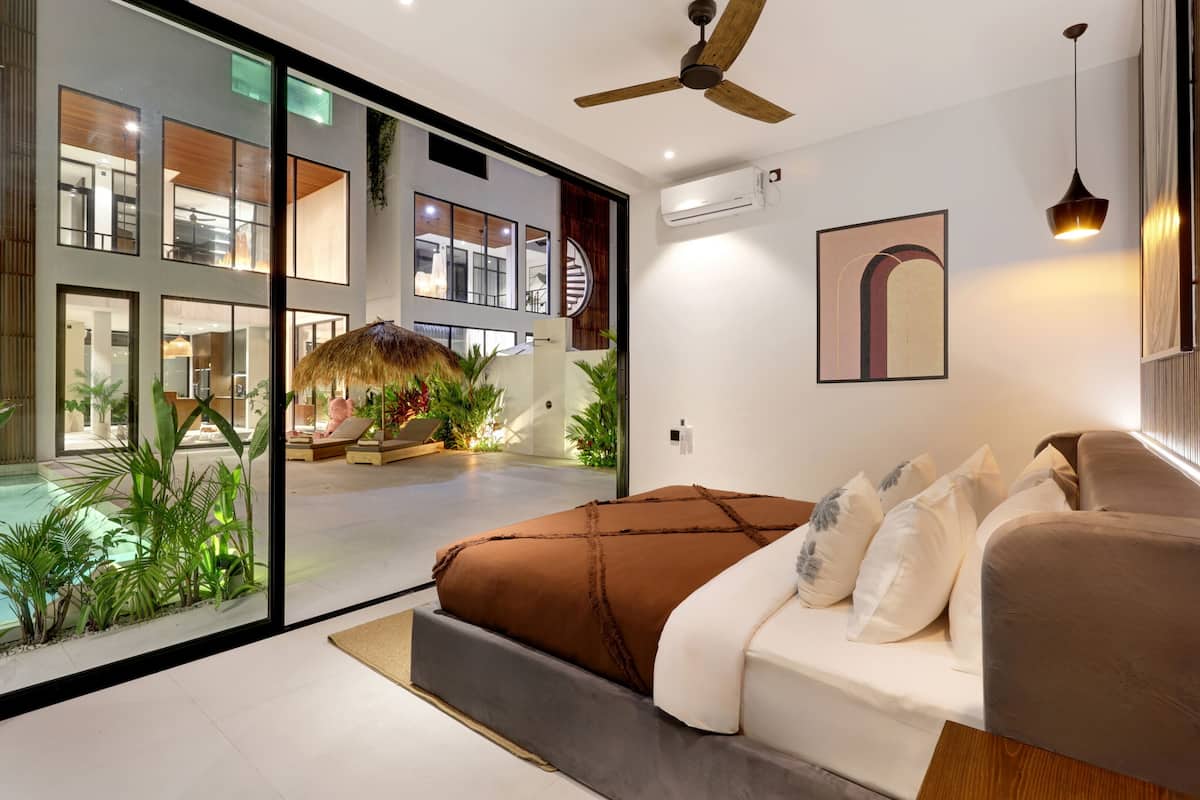 A modern bedroom with a large bed, a ceiling fan, and a view of a pool and outdoor area.