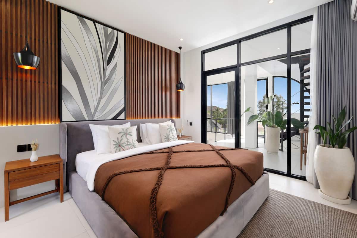 A modern bedroom with a large bed, a nightstand, a wall-mounted light, and a view of the outdoors through a sliding glass door.