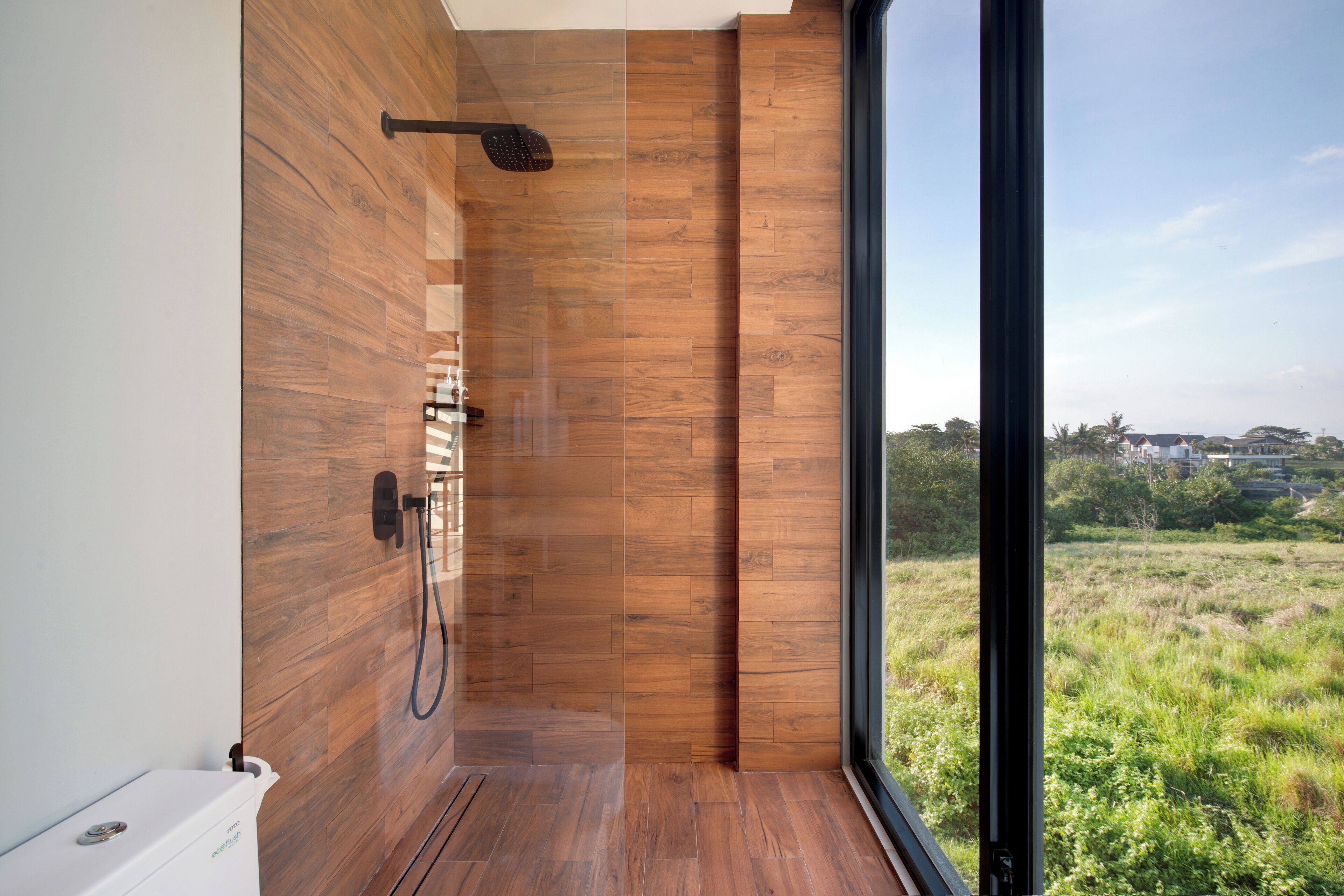 Un baño moderno con suelo de madera y un amplio ventanal que ofrece vistas a un campo cubierto de césped y edificios a lo lejos.
