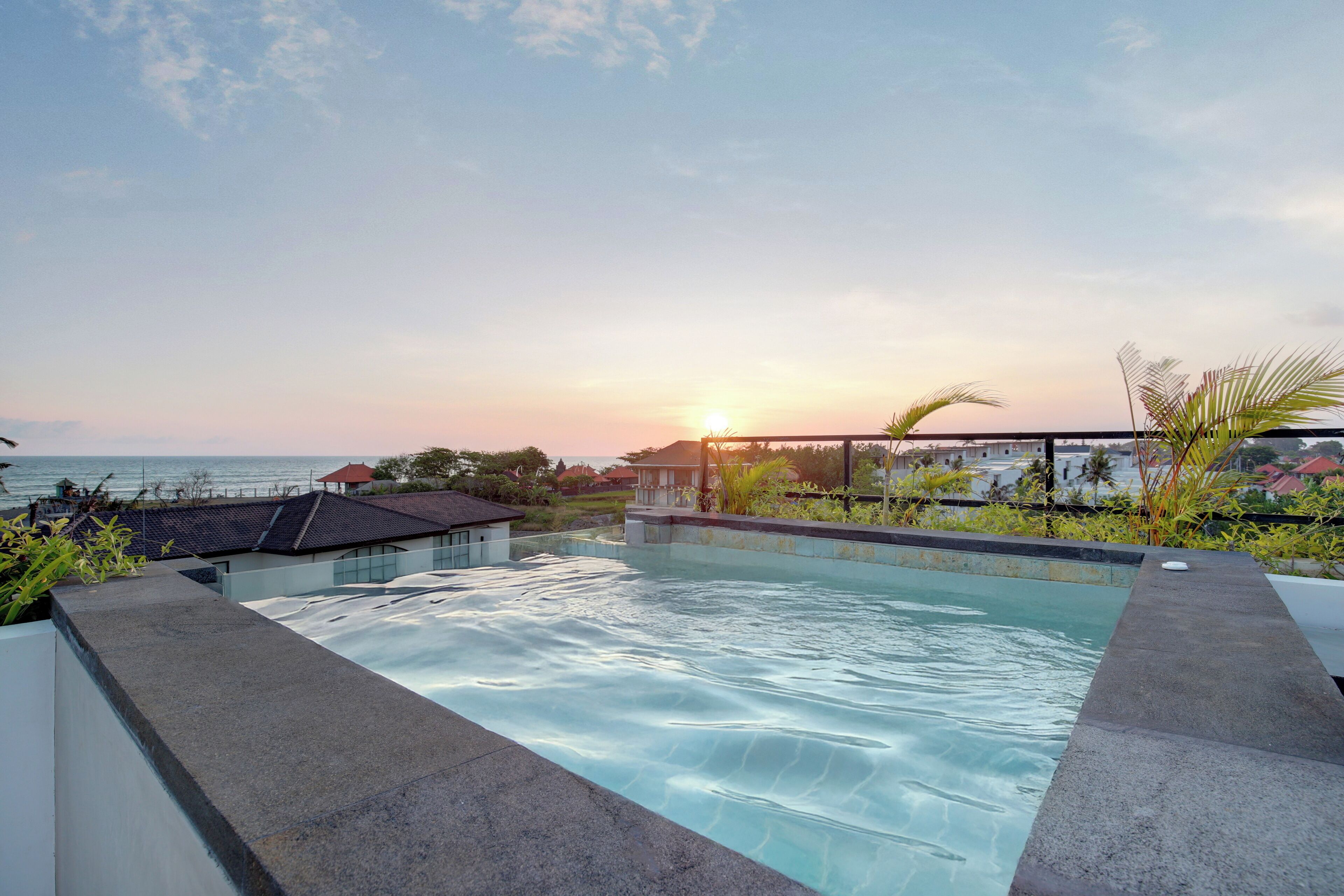 Piscina con vistas al atardecer sobre el océano, rodeada de vegetación y edificios.