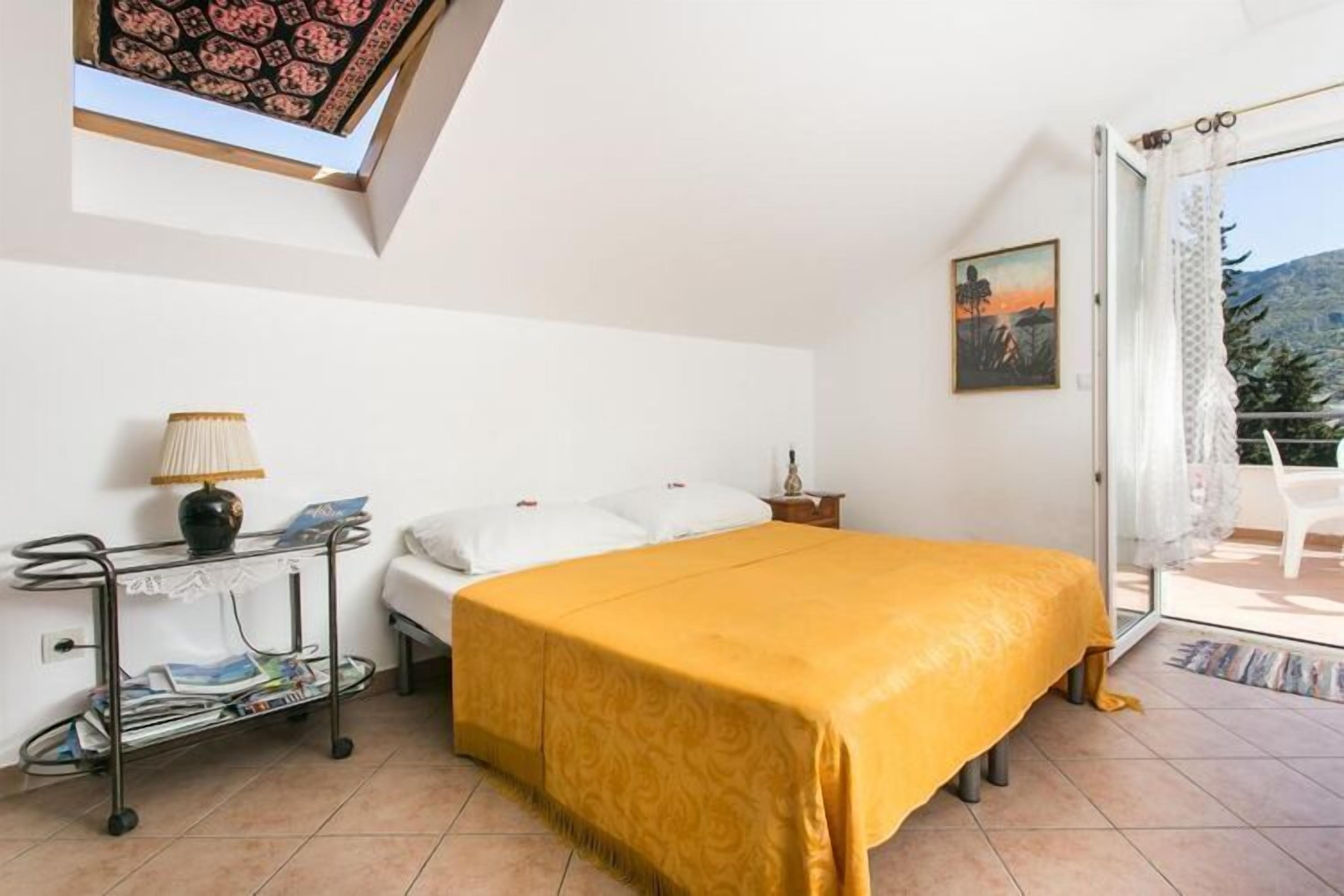 Ein Schlafzimmer mit einem Bett, einem Nachttisch, einer Lampe, einer kleinen Ablagefläche und Blick ins Freie durch eine Glastür.