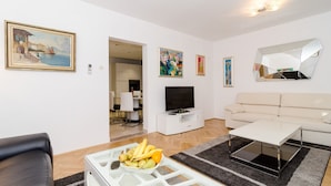 Una sala de estar moderna con un sofá blanco, una mesa de centro de vidrio con fruta, un televisor de pantalla plana sobre un mueble blanco y un espejo grande en la pared.