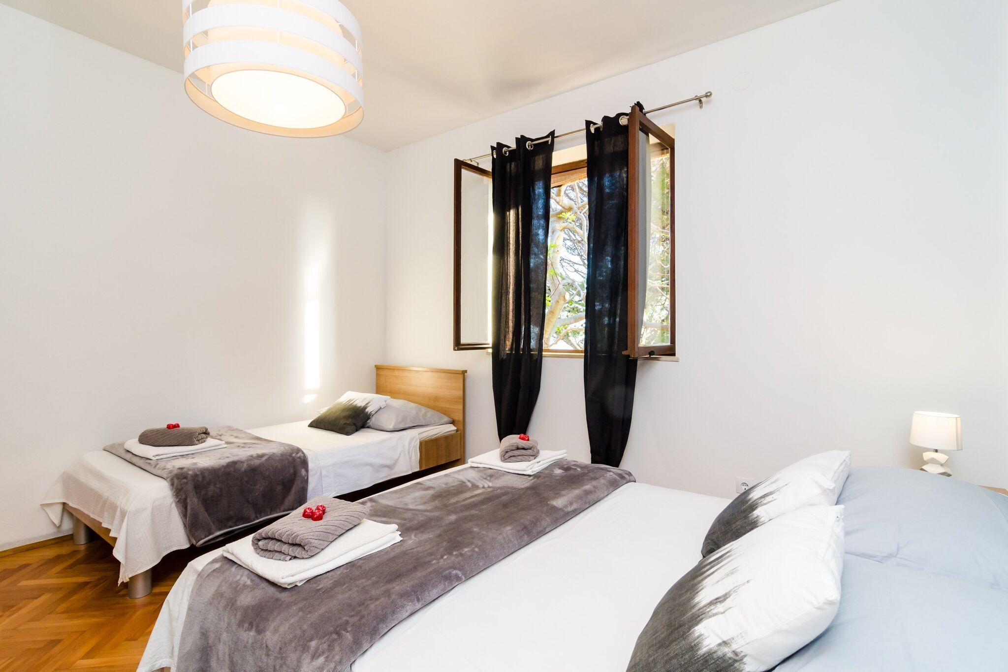 A hotel room with two single beds, a large bed, a window with curtains, a hanging light fixture, and a bedside table with a lamp.