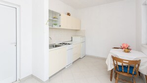 Cocina compacta con armarios blancos, estufa y lavamanos. Hay una mesita con una silla y un florero.