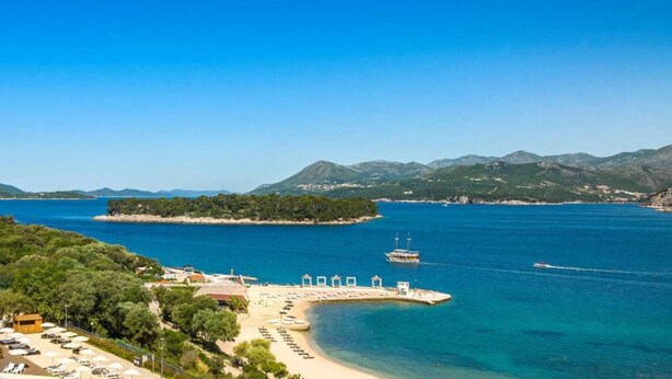 Un hotel frente al mar con playa, muelle para barcos y montañas al fondo.
