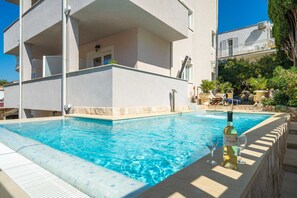 Une piscine, une bouteille de vin et des verres, le tout entouré d’un bâtiment doté de balcons et agrémenté de plantes en pot.
