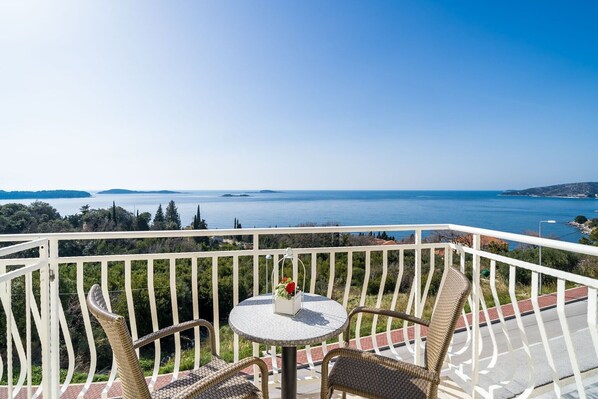 A balcony with wicker chairs, a small round table with a vase of flowers, and a view of the sea and distant islands.