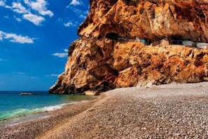 A rocky cliff with a building on top, a pebble beach, and the sea.