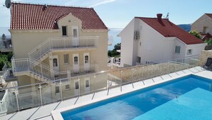 A swimming pool with a glass balustrade, a multi-story building with a red-tiled roof, and a clear view of the sea.