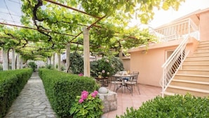 Un chemin de pierre mène à un espace extérieur abrité, aménagé avec une table et des chaises, et entouré de haies soigneusement taillées et d’une pergola couverte de plantes grimpantes.