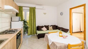 Cocina compacta con estufa a gas, microondas y un refrigerador pequeño. Una mesa de comedor con mantel de encaje y una botella de licor se encuentra en el centro.