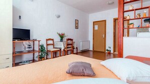 Ein Schlafzimmer mit einem Bett, Kissen, einem Schreibtisch mit Computer, einem Sessel, einem Fernseher und einem Regal mit Dekoration.