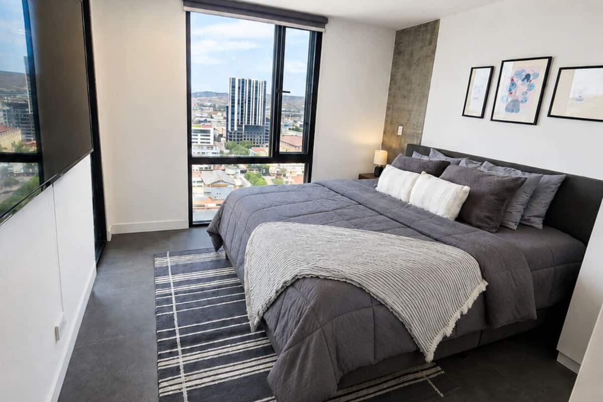 A modern bedroom with a large bed, a view of the city, and a mounted TV.