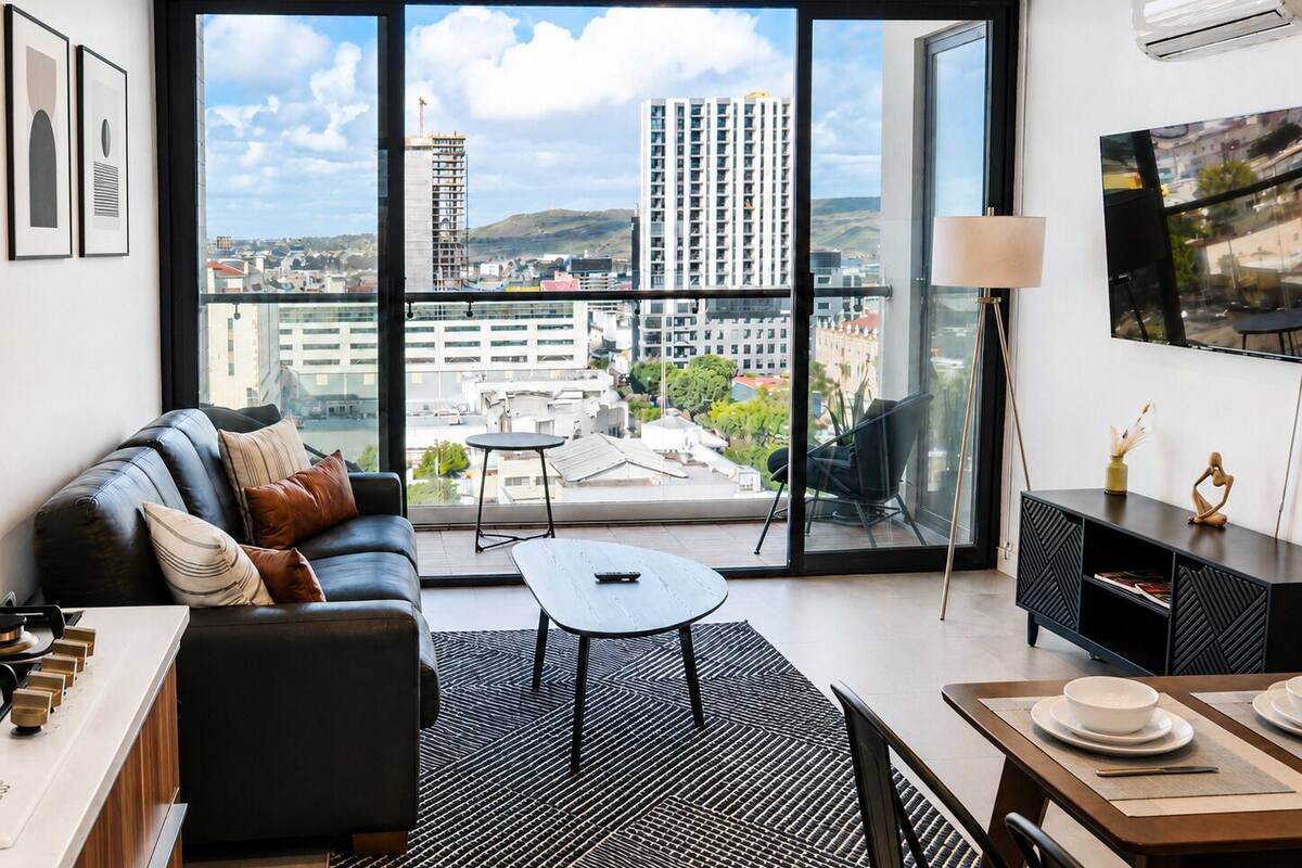 A modern living room with a sofa, coffee table, dining table, and a large window with a city view.