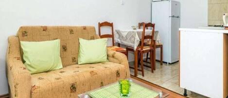 A small, furnished room with a sofa, a dining table, chairs, a kitchen area with a refrigerator, and a microwave.