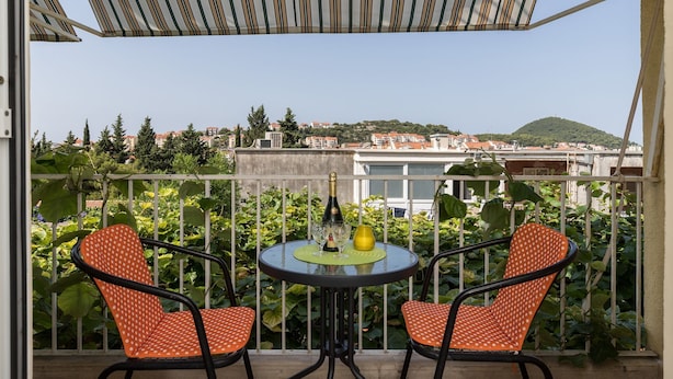 A balcony with two orange chairs, a table with a bottle and glasses, and a view of buildings and greenery.