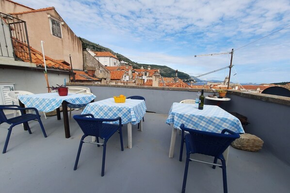 A terrace with checkered tablecloths and blue chairs, overlooking a town with red-roofed buildings.
